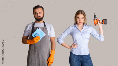 Gender stereotypes and non female profession. Serious man in apron and rubber gloves holding sponges and woman holding a drill photo