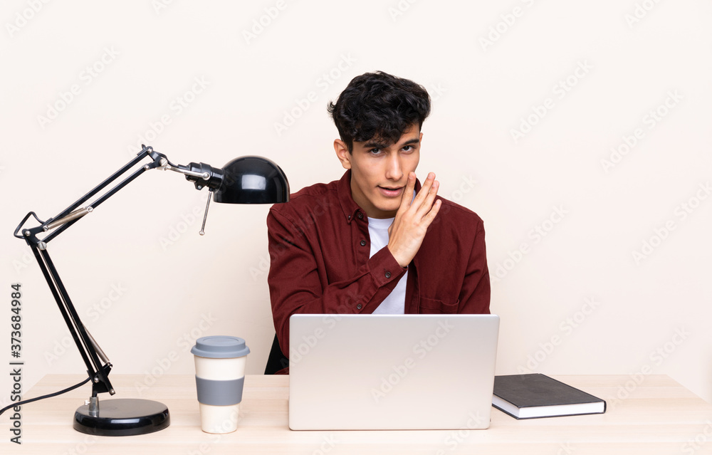 Young Argentinian man in a table with a laptop in his workplace whispering something