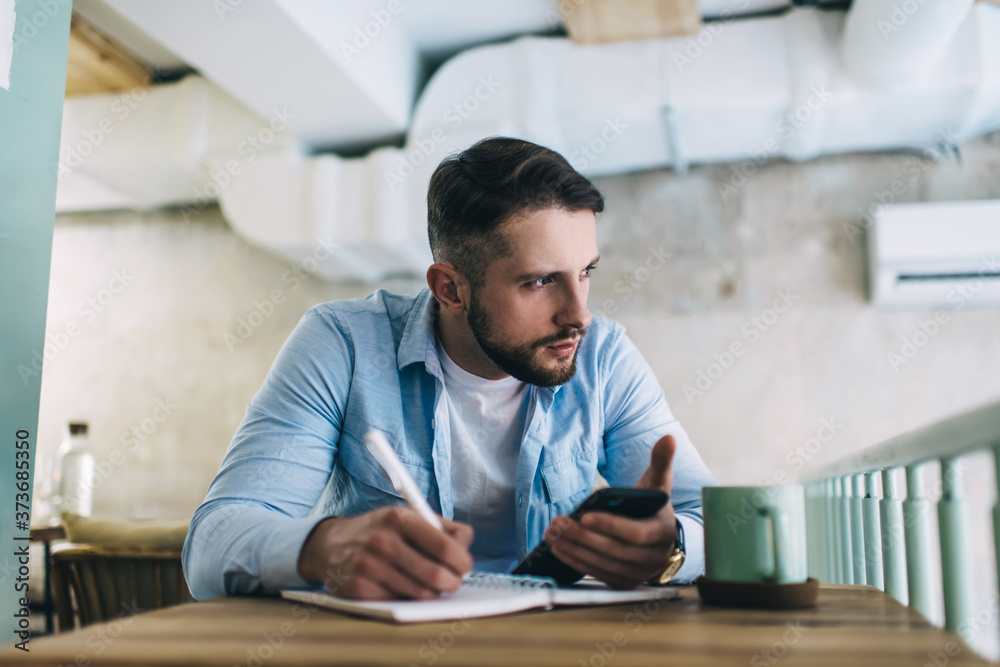 Thoughtful male journalist using smartphone for writing article in notebook while sitting in cafe