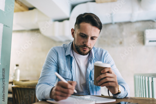 Serious man making notes in notebook while drinking coffee in cafeteria