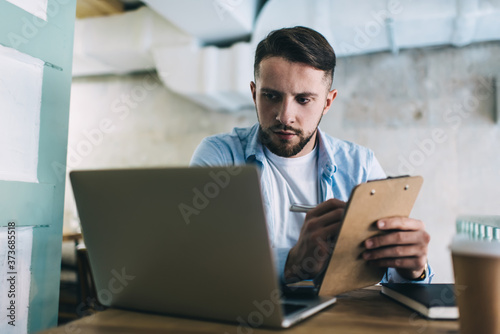 Concerned man making notes in clipboard using laptop