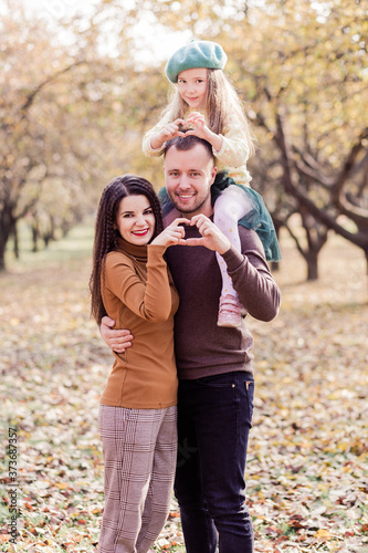 Mom, dad and their little daughter are posing in the autumn park. The girl sits on the shoulders of dad