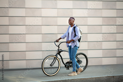 Handsome black man with backpack and bike walking near brick wall outdoors, free space