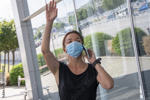 Street portrait of a 35-year-old woman in a medical mask and a phone. Cute brunette waving at someone with your hand. Maybe she saw someone she knew or called a taxi.