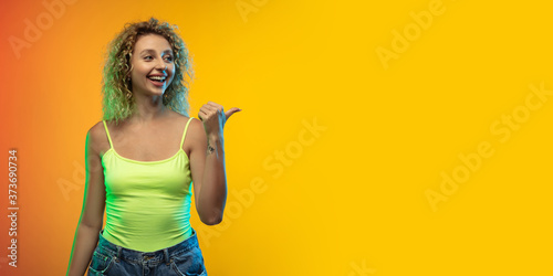 Pointing. Caucasian young woman's portrait on gradient studio background in neon. Beautiful female curly model in casual style. Concept of human emotions, facial expression, youth, sales, ad. Flyer