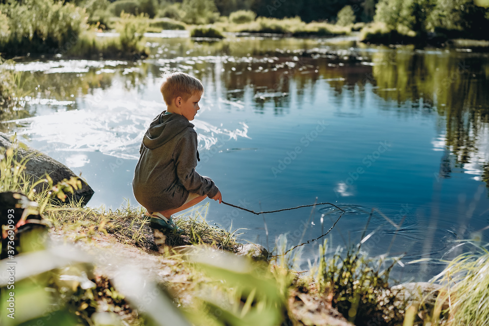 young caucasian boy pretending to do fishing in a pond holding a long rod on sunset. forest and sky reflecting ib water. Image with selective focus young caucasian boy pretending to do fishing in a pond holding a long rod on sunset. forest and sky reflecting ib water. Image with selective focus