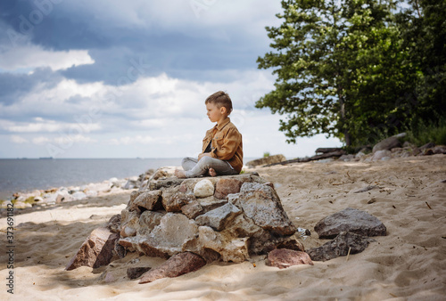 cute caucasian boy sitting on heap big rocks on the coast and meditatively staring at gulf of finland in russia, leningrad region and beautiful cloudy sky