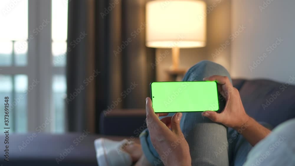 Man at home lying on a sofa and using smartphone with green mock-up screen in horizontal mode. He browsing Internet, watching content, videos, blogs. POV.