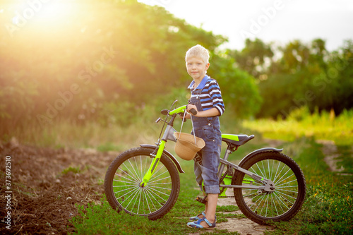happy baby boy blond with a Bicycle stands in a field in summer, children's lifestyle