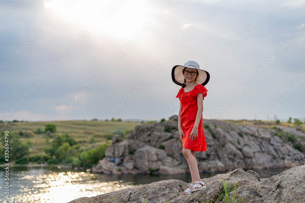 A girl in a red dress and white hat stands on a rock against the backdrop of mountains, a river and a sunset. Happy childhood.