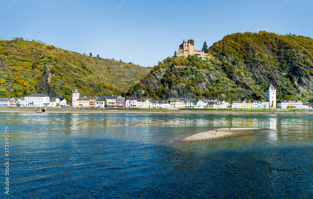 Sunny view of the village Sankt Goarshausen with white houses on the river Rhine in Germany and a castle on a hill above the village.