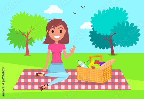 Young girl sitting at checkered plaid in park with basket full of products. Smiling girl relaxing at nature, summer time. Pretty female wearing t-shirt, jeans resting at picnic. Girl show ok sign