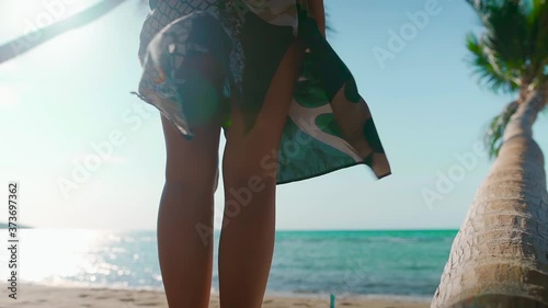 Woman stands on sandy beach with palm tree barefoot wearing light flowing dress blowing in the wind on tropical island