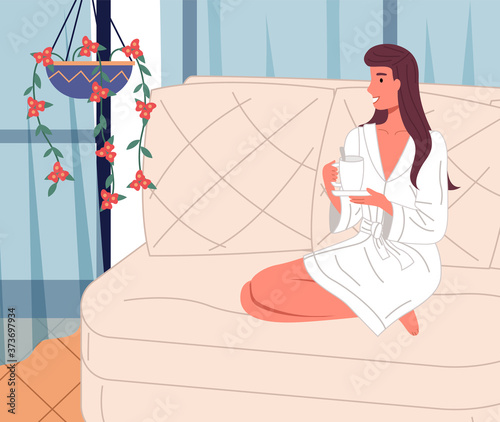Happy smiling woman wearing bathrobe relaxing at sofa with a cup of drink at home. Cheerful brunette looking at curtains and hanging houseplant. Relaxation with tea or coffee after work at home