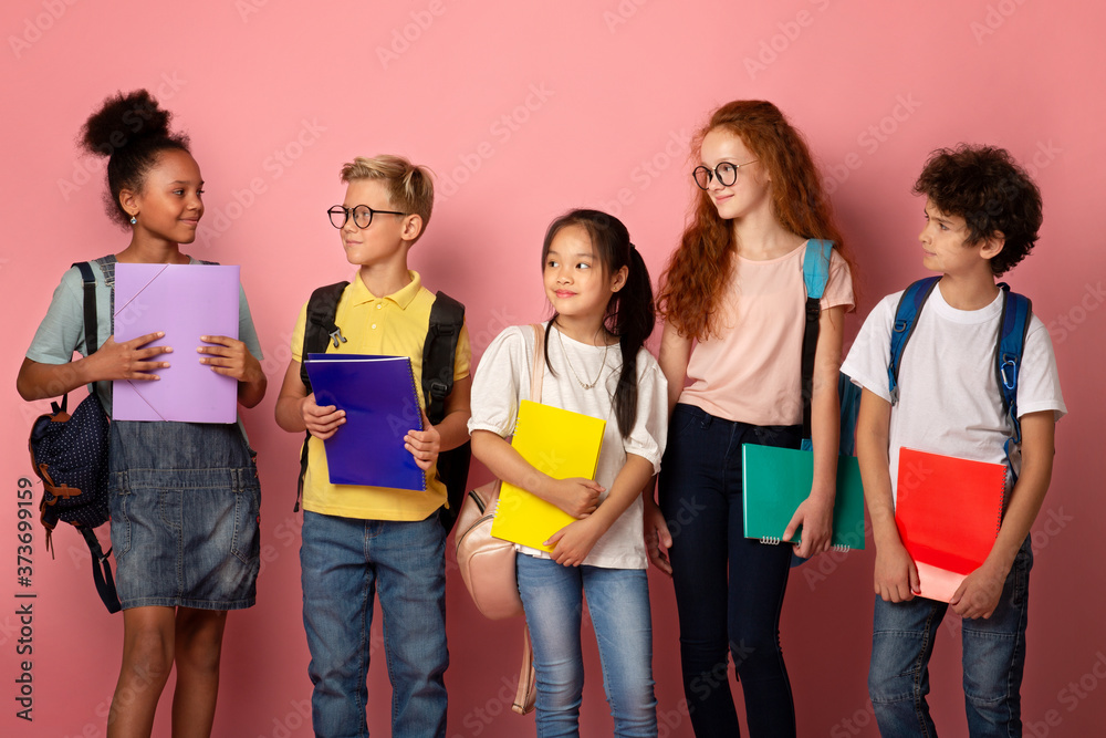 Multinational kids with schoolbags and notebooks looking at each other over pink background Multinational kids with schoolbags and notebooks looking at each other over pink background