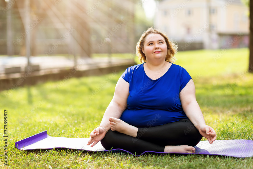Fototapeta premium Young plus size blonde meditates on a gymnastic rug on a sunny summer evening. Healthy lifestyle concept