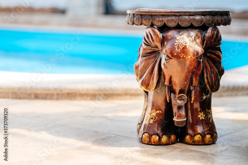 Wooden elephant with artistic engraving stands on the floor near the pool, traditional asian souvenir. Elephant chair