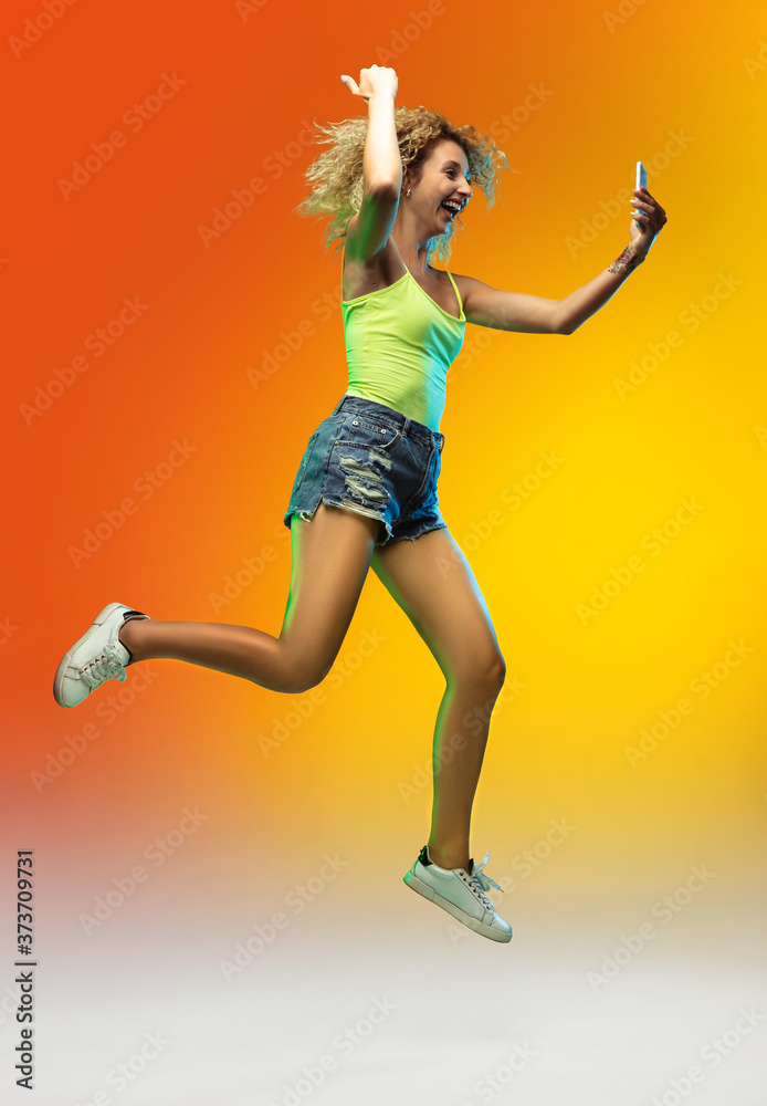 Winner. Jumping caucasian young woman's portrait on gradient studio background in neon. Beautiful female curly model in casual style. Concept of human emotions, facial expression, youth, sales, ad.