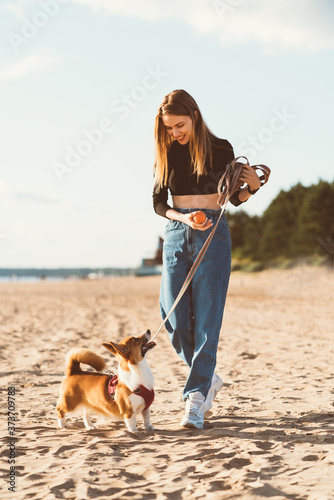 Beautiful young woman playing with Corgi puppy walking along ocean beach. Female looking to dog on Sunny day on ocean coastline. Active lifestyle, pet care
