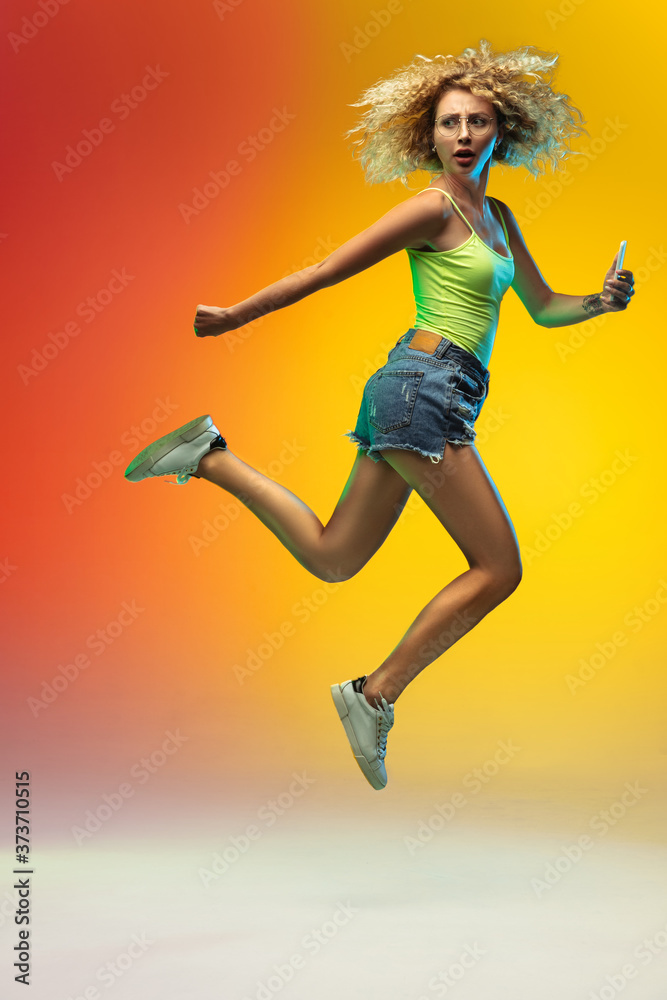 Hurrying up. Jumping caucasian young woman's portrait on gradient studio background in neon. Beautiful female curly model in casual style. Concept of human emotions, facial expression, youth, sales