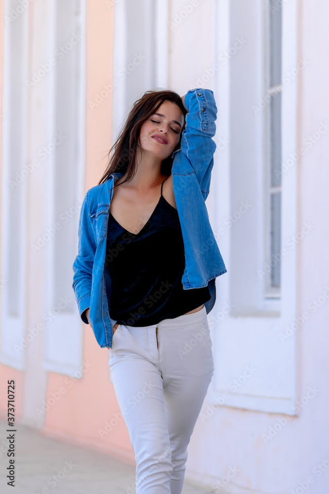 Girl posing in light hall. Hand over head, closed eyes. Casual clothes. White jeans, black top and blue shirt. Blurred background. Girl posing in light hall. Hand over head, closed eyes. Casual clothes. White jeans, black top and blue shirt. Blurred background.