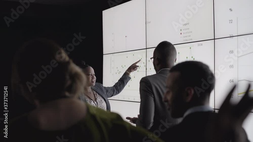 Rear view of a diverse group of businesspeople
watching a presentation by two African
business people on a digital multi screen wall in a dark
office photo