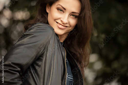 Beautiful brunette smiling business successful woman enjoying in leather black jacket doing active joyful moving on summer day trees colors. Closeup