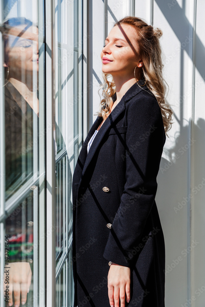 A blonde girl in a black suit stands near the window and enjoys the sunlight