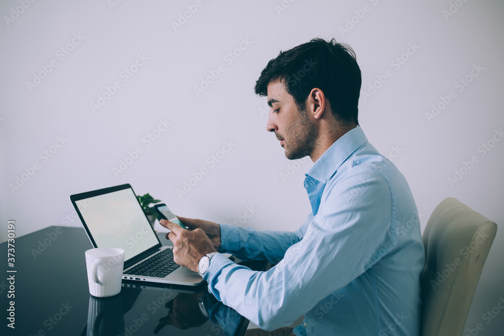 Serious adult executive man using smartphone while sitting at laptop with mug of hot drink Serious adult executive man using smartphone while sitting at laptop with mug of hot drink