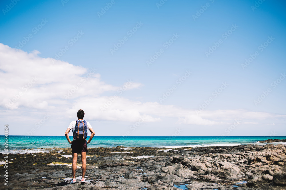 Back view of young backpacker adoring beauty of nature landscape at Menorca coastline, male tourist with travel rucksack spending summer vacations for visiting paradise environment for recreate