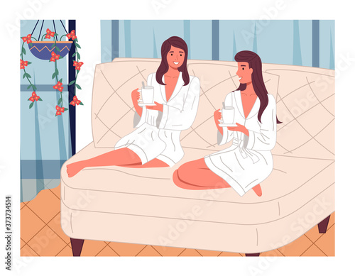 Two young beautiful smiling women are sitting with bent legs on a large bright comfortable sofa with cups of tea or drink, white bathrobes. Girls in the spa. Beauty treatments and relaxation