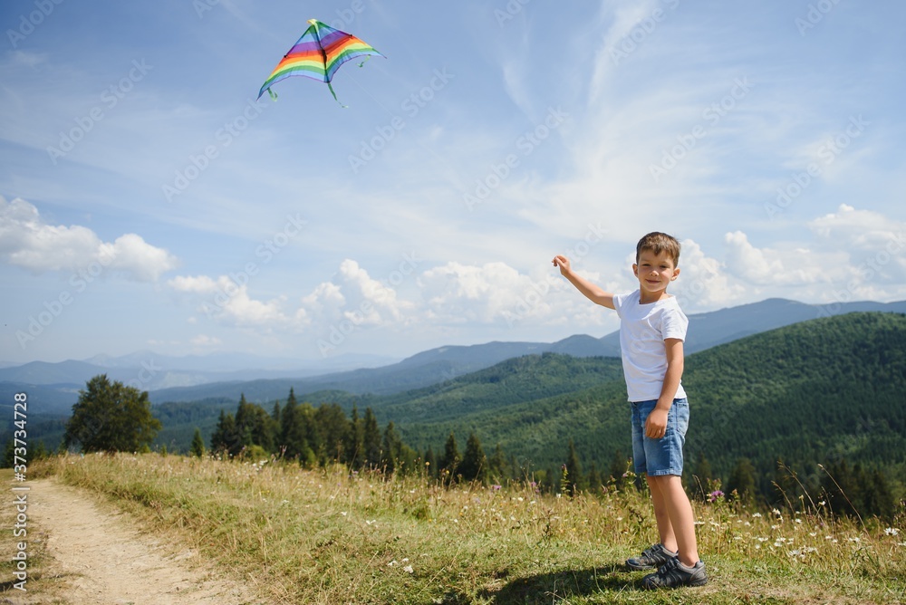 Fototapeta premium Little school age boy running down the slope with kite in the sun. Sunny summer or spring day at sunset. Active outdoor games and leisure. Mountain landscape with hills and coniferous trees.