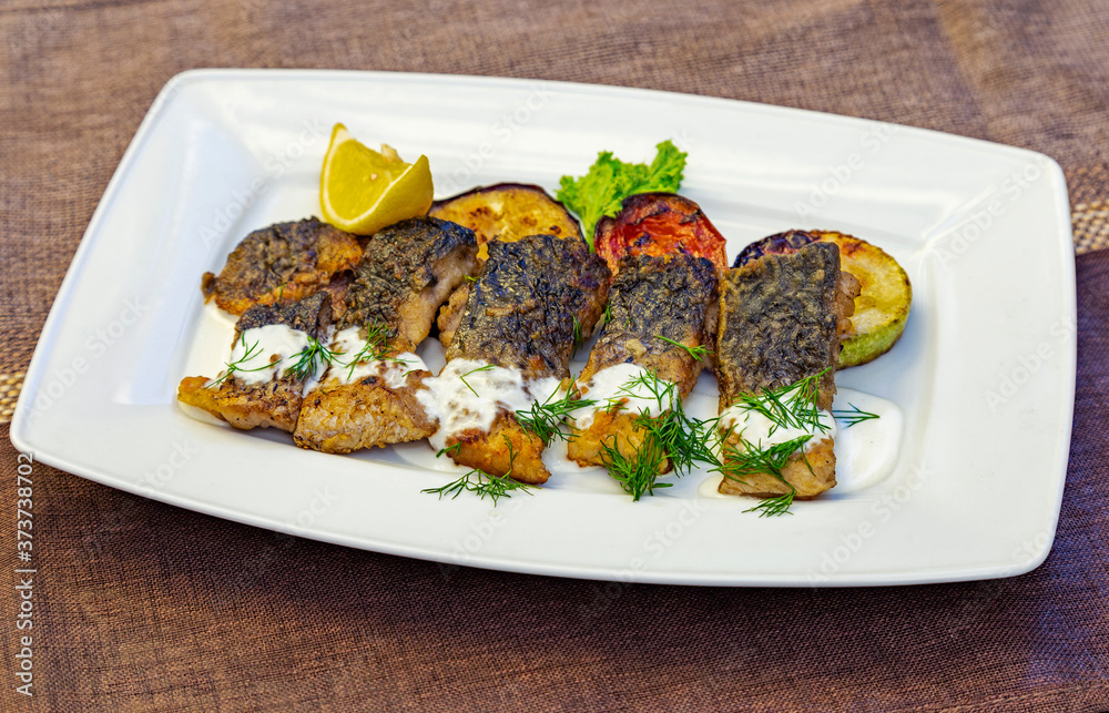 
Pieces of fried fish fillet with grilled vegetables (zucchini, tomato) decorated with lemon, dill and lettuce sprinkled with sour cream sauce. Fish on a white rectangular plate