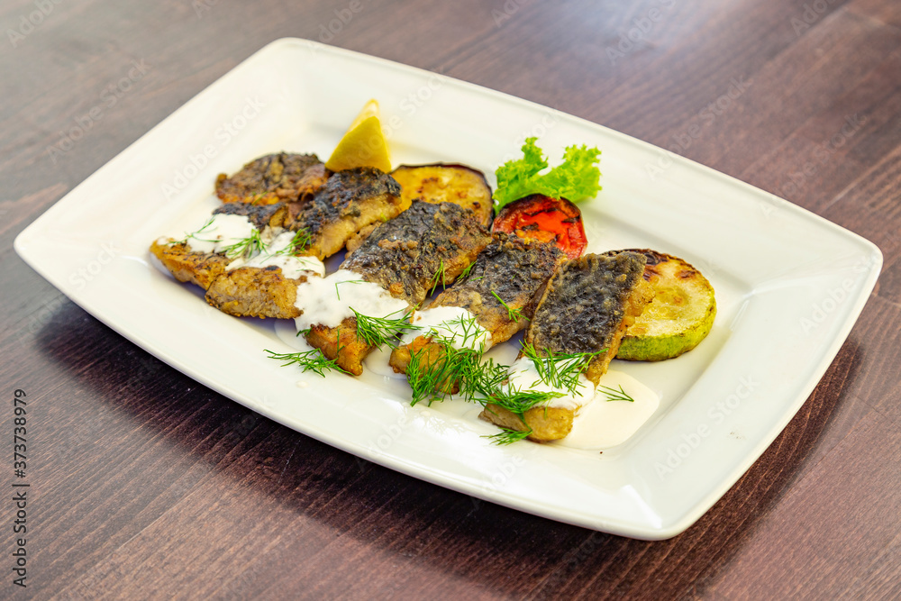Pieces of fried fish fillet with grilled vegetables (zucchini, tomato) decorated with lemon, dill and lettuce sprinkled with sour cream sauce. Fish on a white rectangular plate
Pieces of fried fish fillet with grilled vegetables (zucchini, tomato) decorated with lemon, dill and lettuce sprinkled with sour cream sauce. Fish on a white rectangular plate