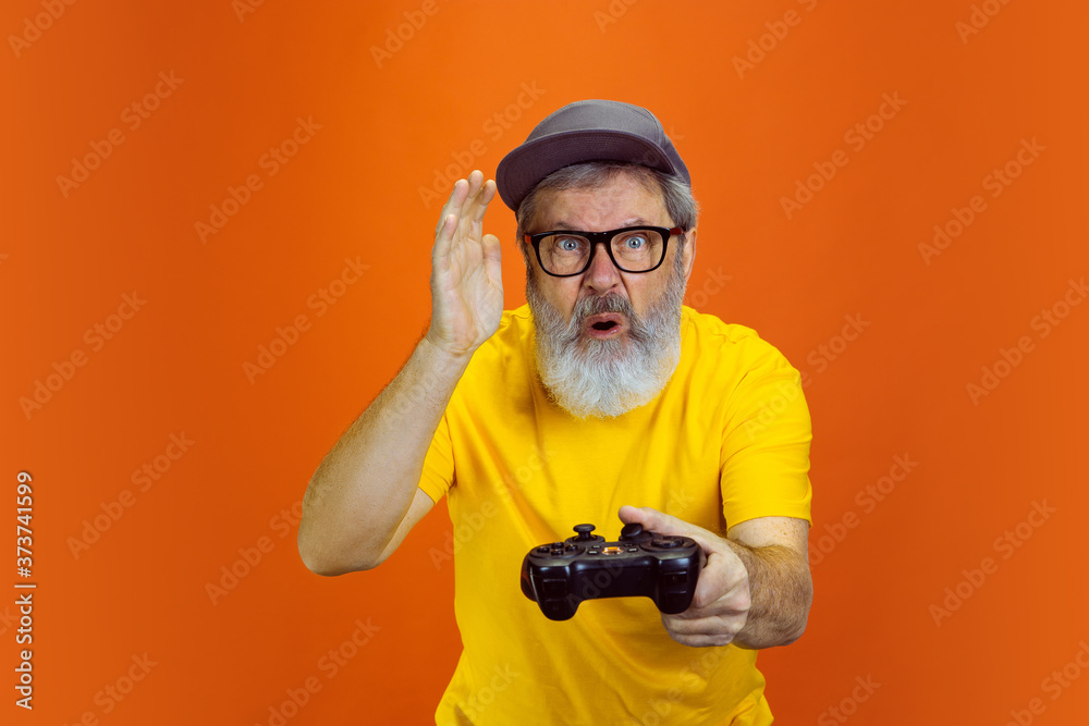 Tensioned gamer. Portrait of senior hipster man using devices, gadgets isolated on orange studio background. Tech and joyful elderly lifestyle concept. Trendy colors, forever youth. Copyspace for your Tensioned gamer. Portrait of senior hipster man using devices, gadgets isolated on orange studio background. Tech and joyful elderly lifestyle concept. Trendy colors, forever youth. Copyspace for your