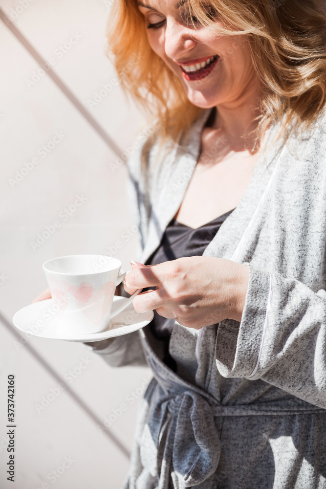 A woman in a silver dressing gown stands near the window with a cup of drink in her hands. The weather is good outside, the sun is shining A woman in a silver dressing gown stands near the window with a cup of drink in her hands. The weather is good outside, the sun is shining