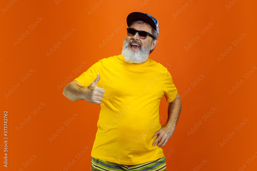 Stunning posing, thumb up. Portrait of senior hipster man in eyewear isolated on orange studio background. Tech and joyful elderly lifestyle concept. Trendy colors, forever youth. Copyspace for your Stunning posing, thumb up. Portrait of senior hipster man in eyewear isolated on orange studio background. Tech and joyful elderly lifestyle concept. Trendy colors, forever youth. Copyspace for your