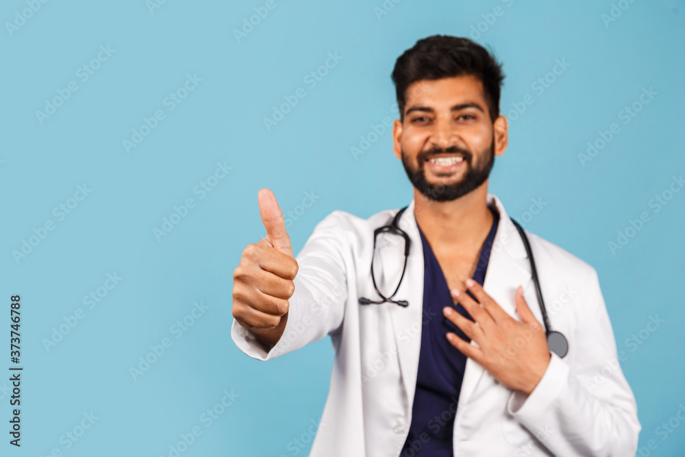 young Indian / Asian doctor with stethoscope in white coat showing thumb up on blue background. young Indian / Asian doctor with stethoscope in white coat showing thumb up on blue background.
