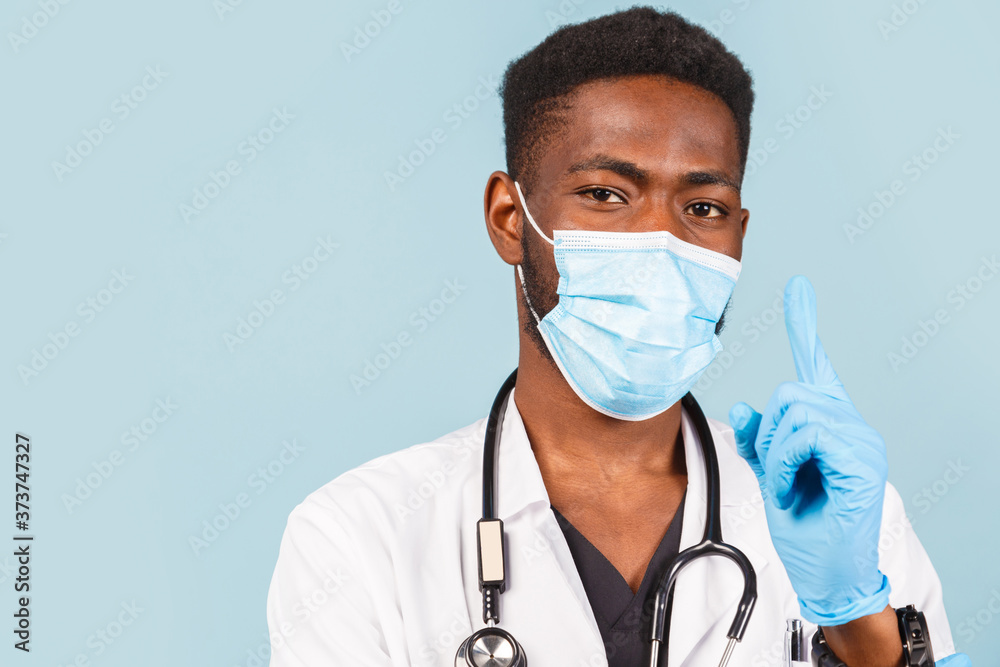 african american male doctor with stethoscope wearing mask and gloves raised his index finger up against blue background. african american male doctor with stethoscope wearing mask and gloves raised his index finger up against blue background.