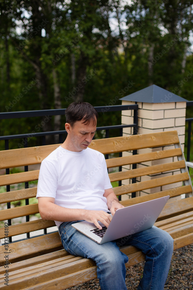 Fototapeta premium Portrait of a middle-aged man with laptop sitting on the bench. Man in white t-shirt and blue jeans with laptop sitting on the bench in the park.