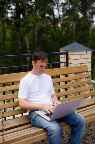 Portrait of a middle-aged man with laptop sitting on the bench. Man in white t-shirt and blue jeans with laptop sitting on the bench in the park.