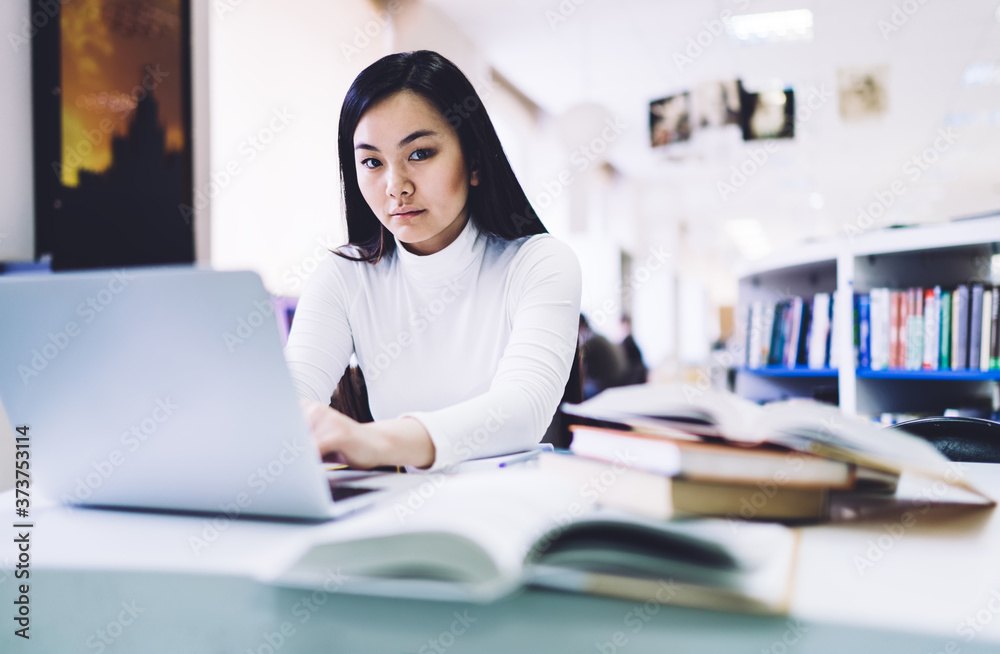 Adult ethnic woman looking at camera while working at laptop in library Adult ethnic woman looking at camera while working at laptop in library