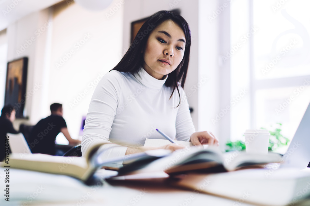 Ethnic woman reading book while sitting at desk and taking notes in notepad in office Ethnic woman reading book while sitting at desk and taking notes in notepad in office