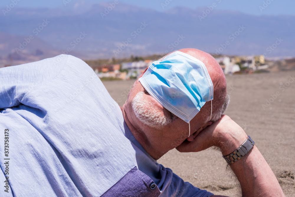 Senior man lying on the sand plays with the coronavirus protective mask putting it on the eyes Senior man lying on the sand plays with the coronavirus protective mask putting it on the eyes