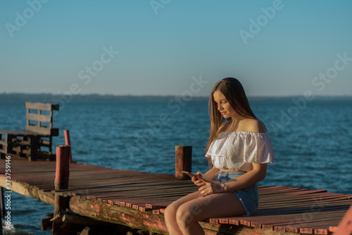 young blonde woman sitting on a pier using her cell phone