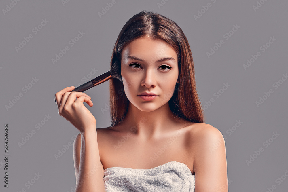 Cute attractive girl with a towel uses a brush for powder or blush isolated on a gray background. Skin care concept. Spa treatments, cosmetology, make-up. Cute attractive girl with a towel uses a brush for powder or blush isolated on a gray background. Skin care concept. Spa treatments, cosmetology, make-up.