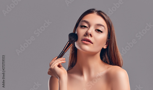 Cute attractive girl with a towel uses a brush for powder or blush isolated on a gray background. Skin care concept. Spa treatments, cosmetology, make-up.