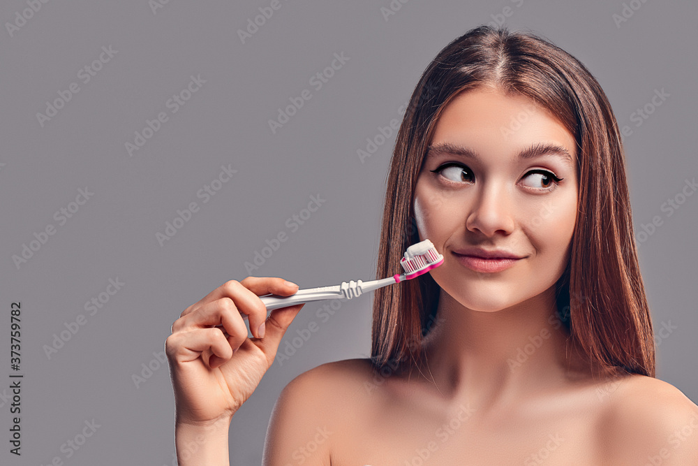 Young attractive brunette girl with loose hair uses a toothbrush and paste isolated on a gray background. Young attractive brunette girl with loose hair uses a toothbrush and paste isolated on a gray background.