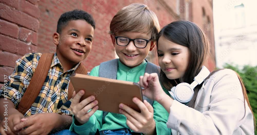 Close up portrait of mixed-races pupils playing on tablet outdoor near school. Happy Caucasian boy and girl tapping and scrolling on device. African American kid talking at schoolyard. Friends concept