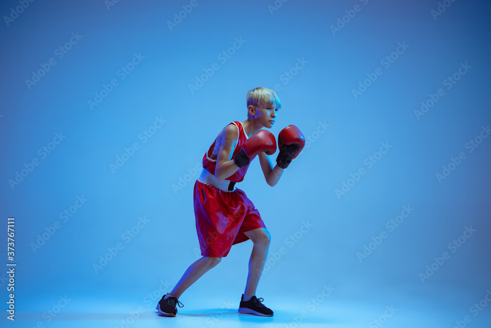 Teenager in sportswear boxing isolated on blue studio background in neon light. Novice male caucasian boxer training hard and working out, kicking. Sport, healthy lifestyle, movement concept.